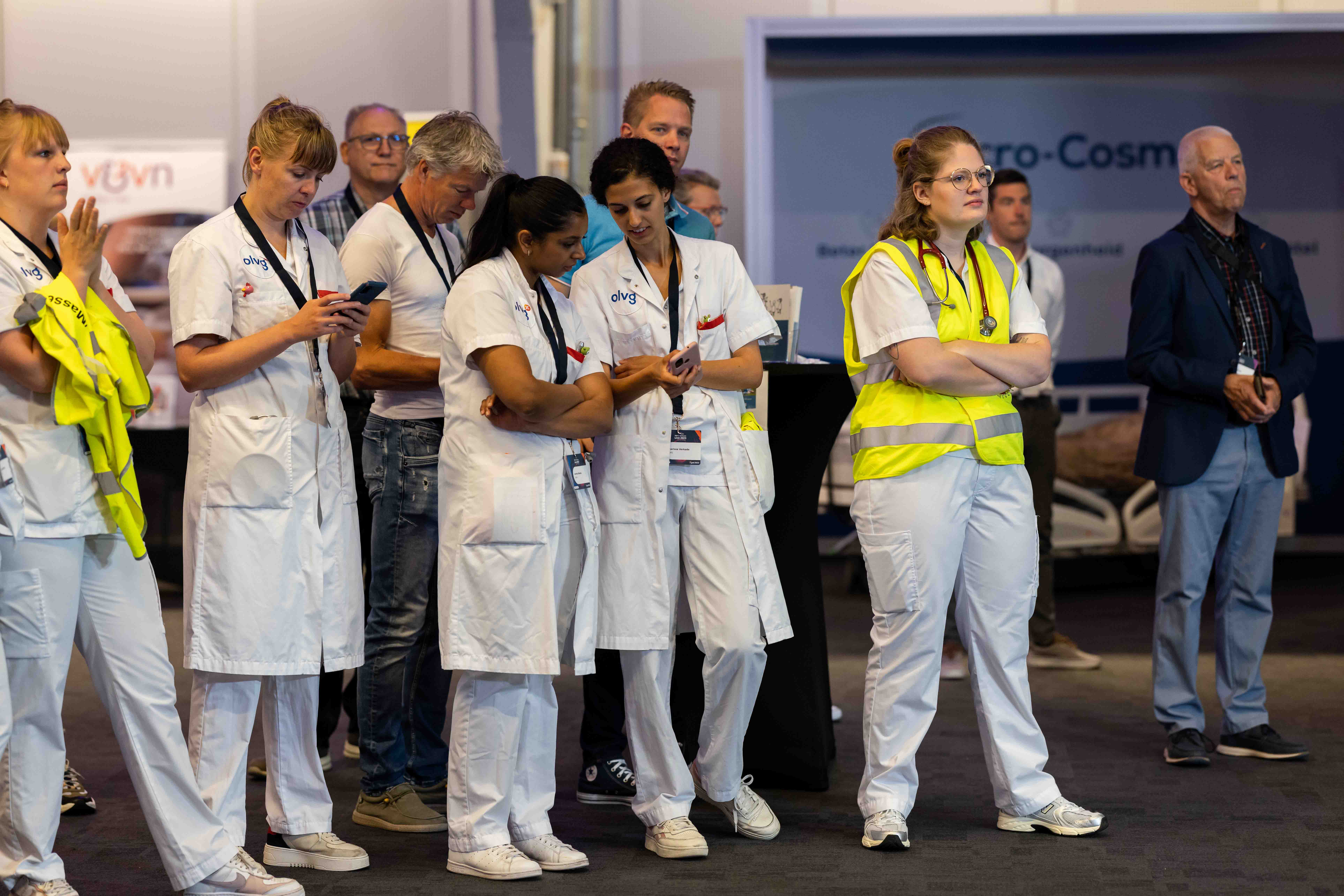 Een groep medische professionals in witte uniformen en één persoon in een geel veiligheidshesje staan aandachtig te kijken naar iets buiten beeld op een beurs of evenement, met bezoekers en stands op de achtergrond.